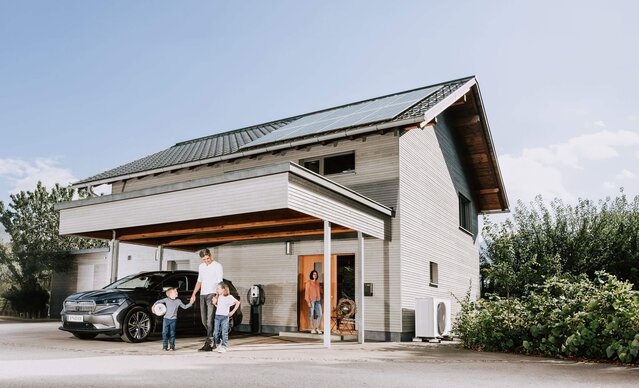 Familie steht vor Einfamilienhaus mit Photovoltaikanlage, Wärmepumpe und Wallbox