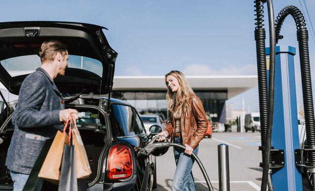 Mann lädt Einkäufe in Kofferraum, während Frau ihr E-Auto ansteckt