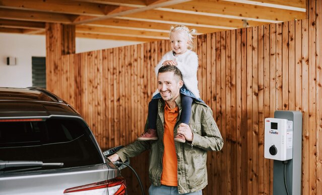 Mädchen sitzt auf den Schultern ihres Vaters, der sein E-Auto auflädt