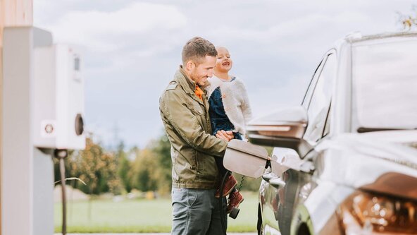Vater mit Tochter im Arm lädt sein E-Auto an einer Wallbox auf